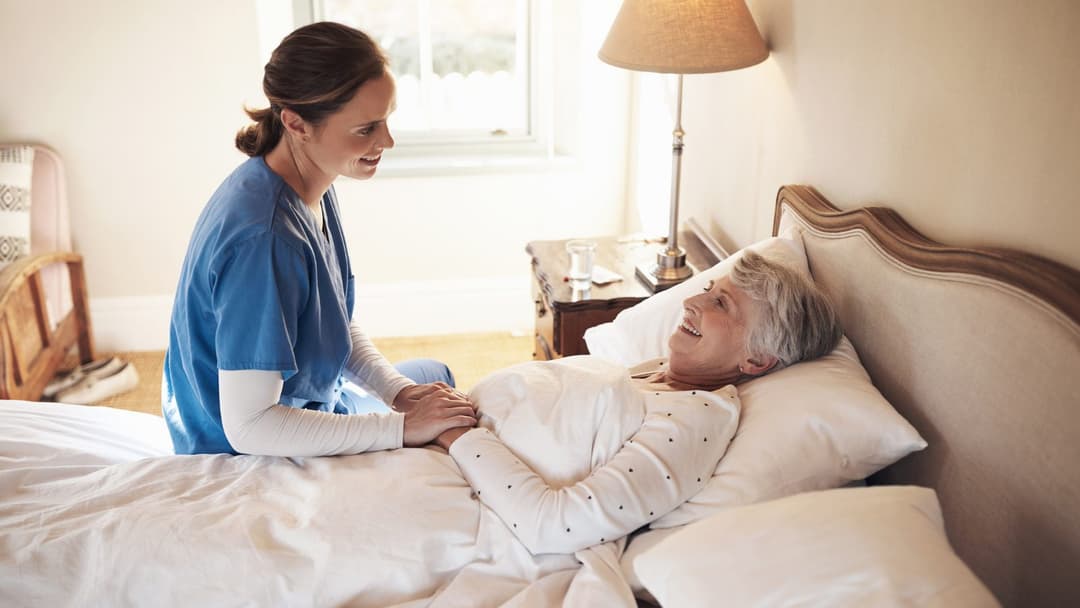 Caregiver in scrubs sitting on the bed and holding the hand of a smiling elderly woman resting in her bedroom