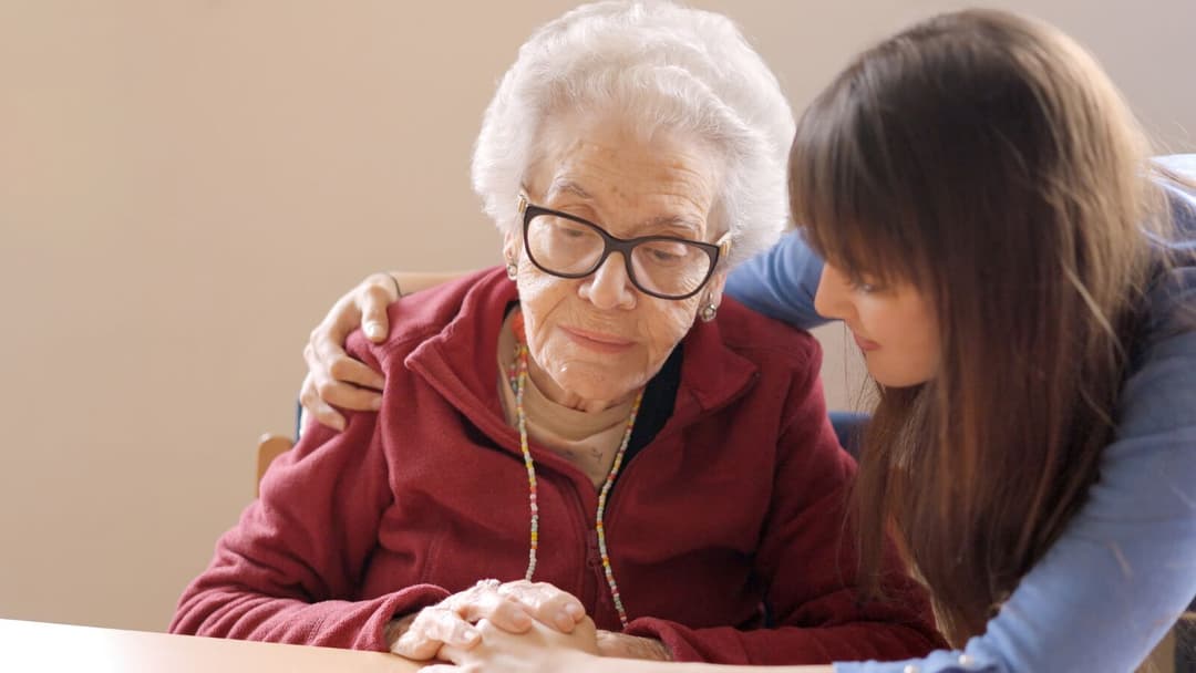 Caregiver with her arm around an elderly woman in glasses and a red sweater, leaning in to talk with her at a table