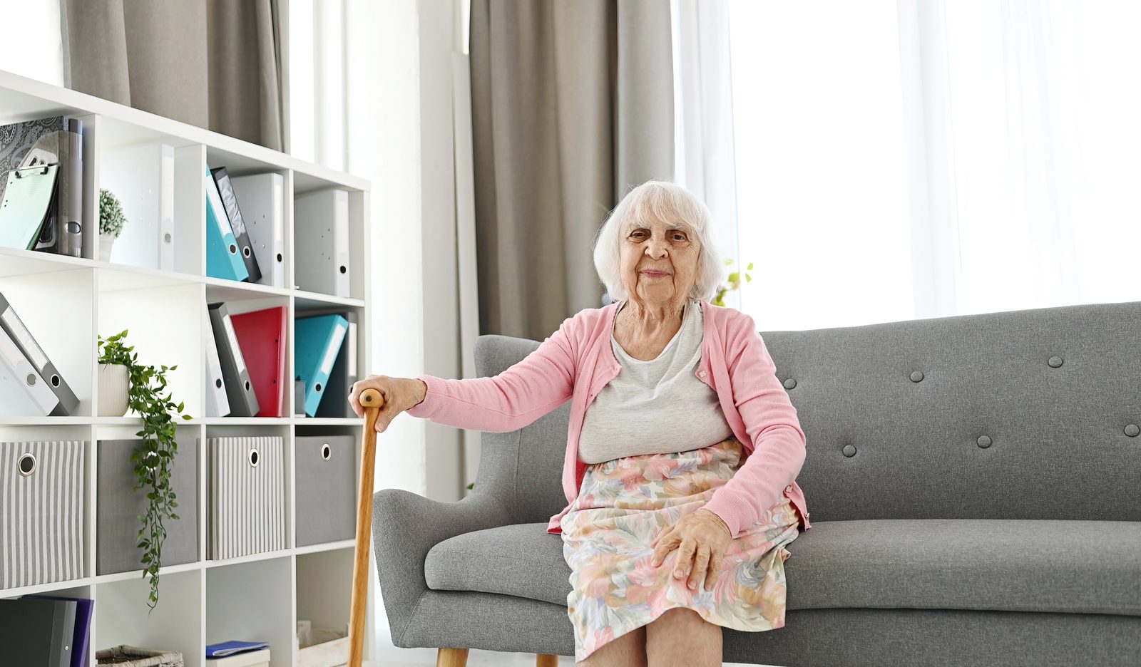 Elderly woman sitting on a living room sofa with her cane, laughing and looking into the camera